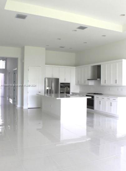 kitchen with white cabinetry, wall chimney range hood, a center island with sink, light tile patterned floors, and appliances with stainless steel finishes