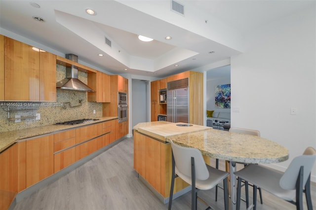 kitchen with light stone countertops, light hardwood / wood-style floors, wall chimney range hood, a tray ceiling, and built in appliances