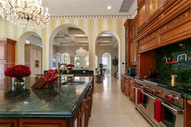 kitchen with coffered ceiling, pendant lighting, range with two ovens, a notable chandelier, and an island with sink