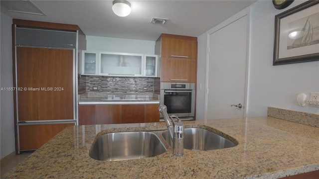 kitchen featuring backsplash, oven, paneled built in refrigerator, light stone counters, and sink