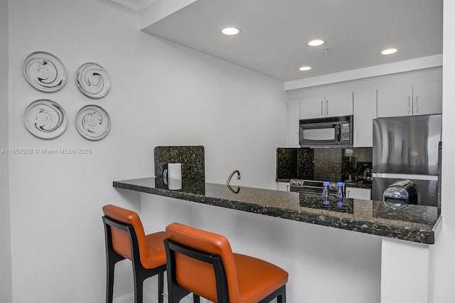 kitchen with white cabinetry, dark stone countertops, a kitchen breakfast bar, stainless steel appliances, and kitchen peninsula