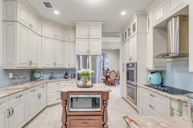 kitchen featuring light stone counters, white cabinets, wall chimney range hood, appliances with stainless steel finishes, and decorative backsplash