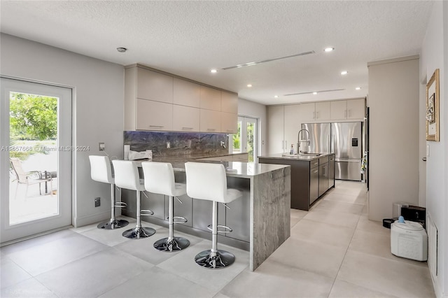 kitchen with a textured ceiling, backsplash, a breakfast bar, stainless steel refrigerator with ice dispenser, and a center island with sink