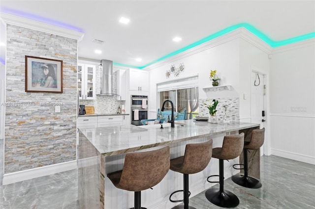 kitchen featuring stainless steel double oven, wall chimney exhaust hood, backsplash, a kitchen bar, and white cabinets