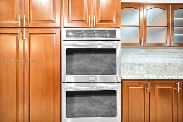 kitchen with backsplash, glass insert cabinets, brown cabinets, light stone countertops, and double oven