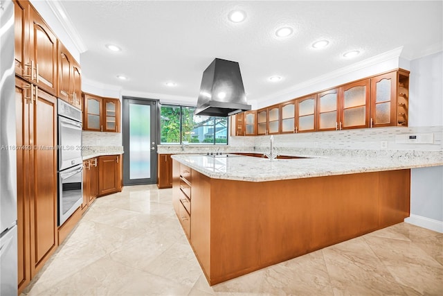 kitchen with light stone counters, island range hood, ornamental molding, a peninsula, and brown cabinetry