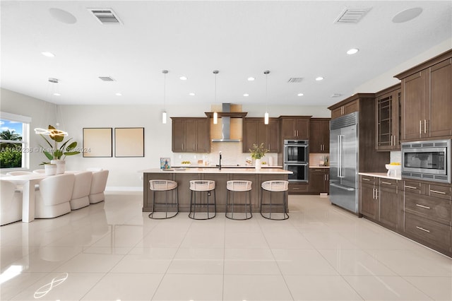 kitchen featuring an island with sink, a kitchen bar, built in appliances, pendant lighting, and dark brown cabinetry