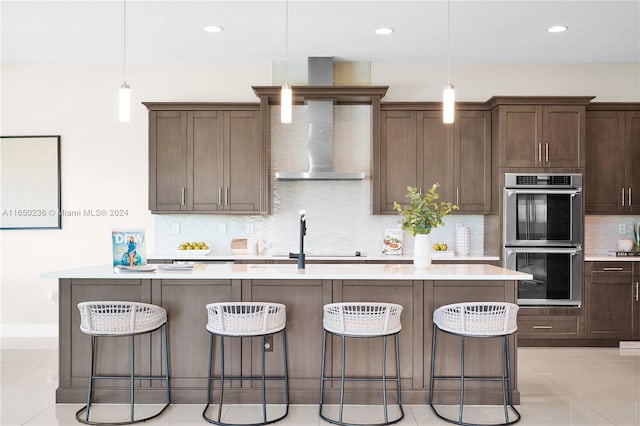 kitchen with hanging light fixtures, stainless steel double oven, an island with sink, and wall chimney range hood