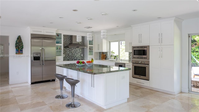 kitchen featuring white cabinets, a kitchen island, wall chimney range hood, dark stone countertops, and built in appliances