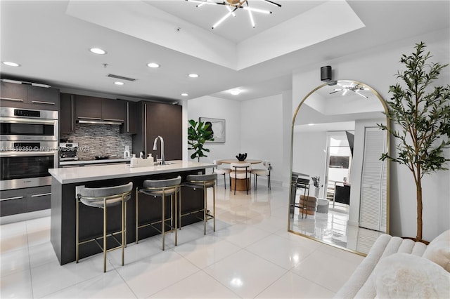 kitchen featuring a tray ceiling, an island with sink, a kitchen breakfast bar, and double oven