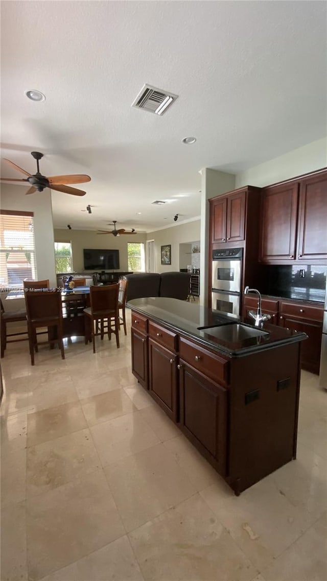 kitchen featuring stainless steel double oven, a kitchen island with sink, sink, ceiling fan, and dark brown cabinetry