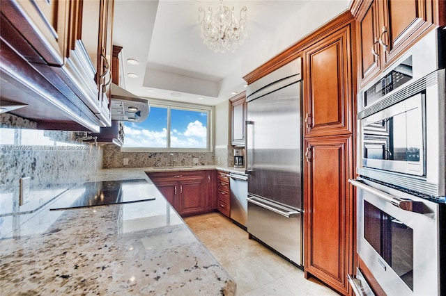 kitchen featuring a notable chandelier, built in appliances, light stone counters, decorative backsplash, and a raised ceiling