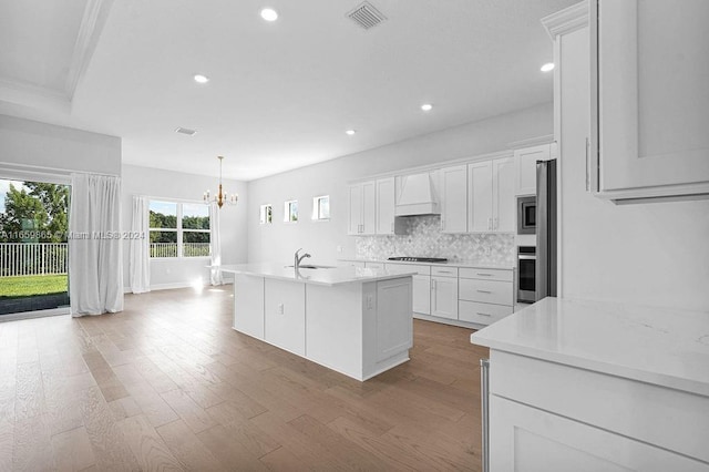 kitchen with white cabinetry, decorative backsplash, a center island with sink, custom exhaust hood, and appliances with stainless steel finishes