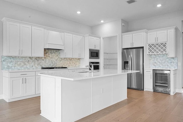 kitchen with white cabinetry, beverage cooler, premium range hood, a kitchen island with sink, and appliances with stainless steel finishes