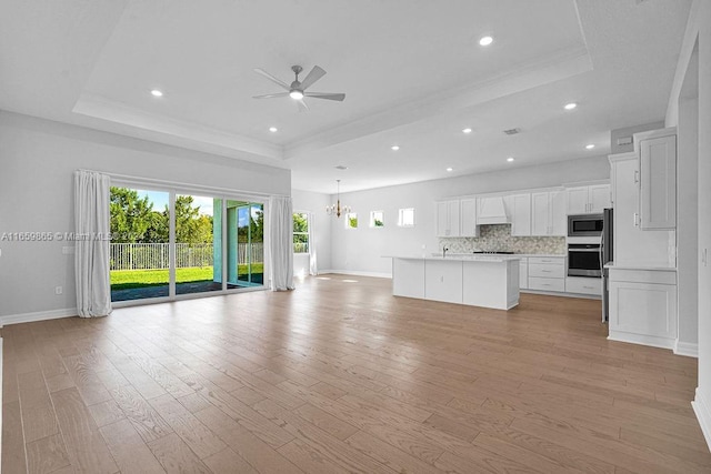 unfurnished living room featuring ceiling fan with notable chandelier, a tray ceiling, and light hardwood / wood-style floors