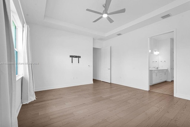 unfurnished bedroom featuring ensuite bathroom, a raised ceiling, ceiling fan, and light hardwood / wood-style flooring