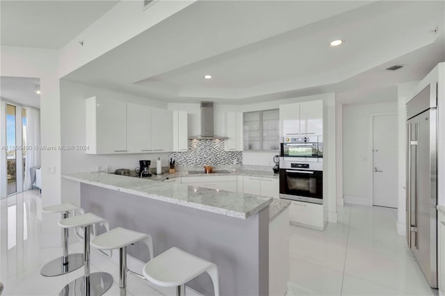 kitchen featuring a breakfast bar area, wall chimney exhaust hood, kitchen peninsula, white cabinetry, and stainless steel double oven