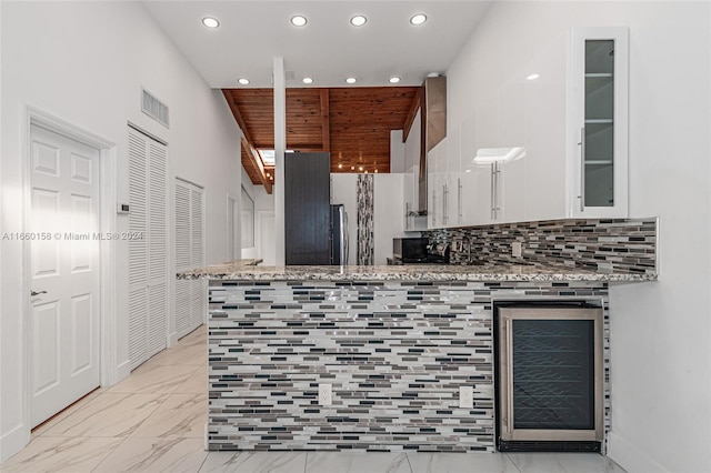 kitchen featuring white cabinetry, backsplash, beverage cooler, kitchen peninsula, and light stone countertops