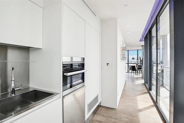 kitchen with a wall of windows, white cabinets, stainless steel oven, and light hardwood / wood-style flooring