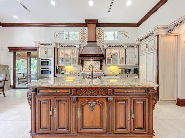 kitchen featuring stainless steel double oven, light stone counters, premium range hood, a large island with sink, and ornamental molding