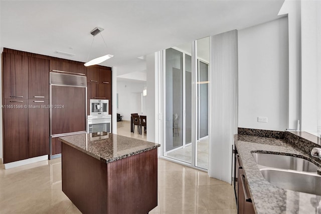 kitchen featuring built in appliances, sink, hanging light fixtures, a kitchen island, and dark stone countertops