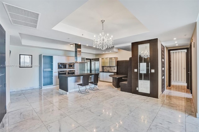 kitchen with a raised ceiling, a kitchen island, a notable chandelier, range hood, and a kitchen bar