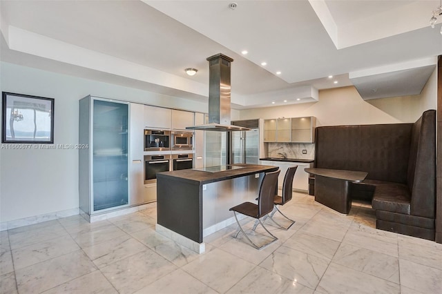 kitchen featuring a tray ceiling, built in appliances, a breakfast bar area, island range hood, and tasteful backsplash