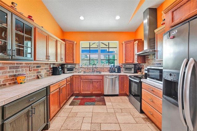 kitchen with sink, wall chimney exhaust hood, stainless steel appliances, tile countertops, and decorative backsplash