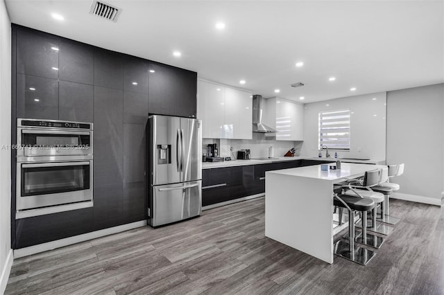 kitchen with a breakfast bar, stainless steel appliances, wall chimney range hood, white cabinetry, and a kitchen island