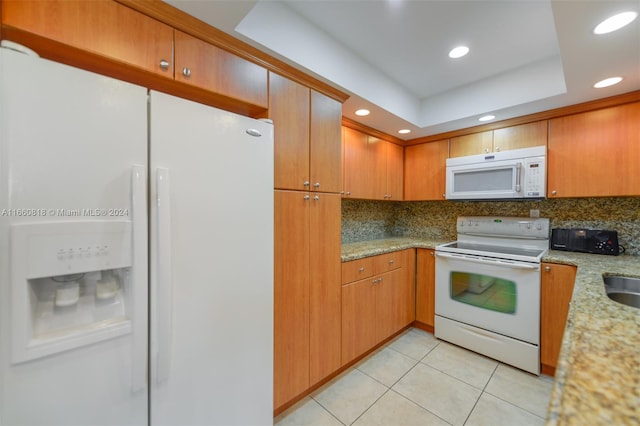 kitchen with a tray ceiling, light stone counters, light tile patterned flooring, decorative backsplash, and white appliances