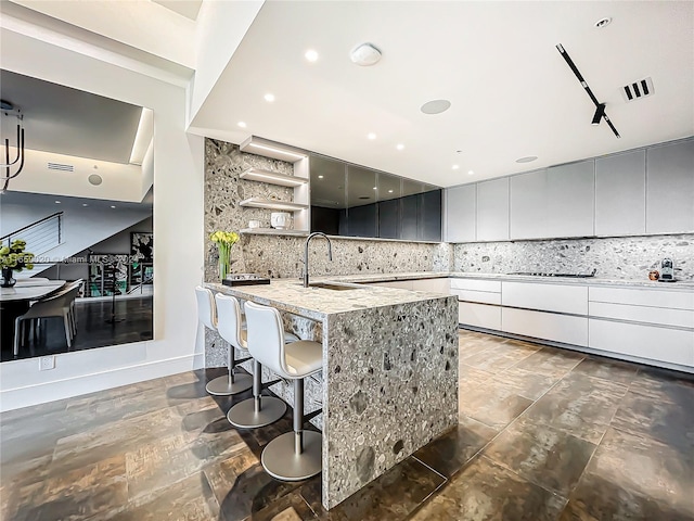 kitchen with an island with sink, backsplash, a breakfast bar area, gray cabinets, and sink