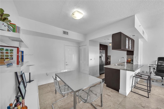 kitchen with dark brown cabinets, a textured ceiling, stainless steel fridge with ice dispenser, a breakfast bar area, and sink