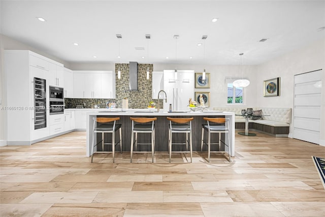 kitchen with white cabinets, an island with sink, hanging light fixtures, wall chimney range hood, and a breakfast bar