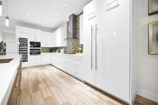 kitchen with decorative light fixtures, backsplash, wall chimney range hood, white cabinetry, and light wood-type flooring