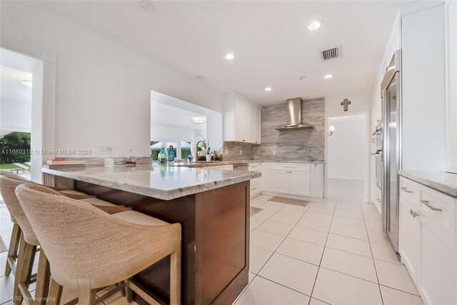 kitchen featuring a kitchen breakfast bar, white cabinetry, kitchen peninsula, and wall chimney range hood