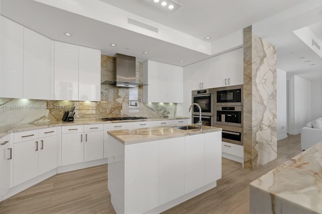 kitchen featuring white cabinetry, wall chimney exhaust hood, light wood-type flooring, a center island with sink, and sink