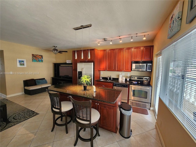 kitchen with ceiling fan, hanging light fixtures, a kitchen island, appliances with stainless steel finishes, and dark stone counters