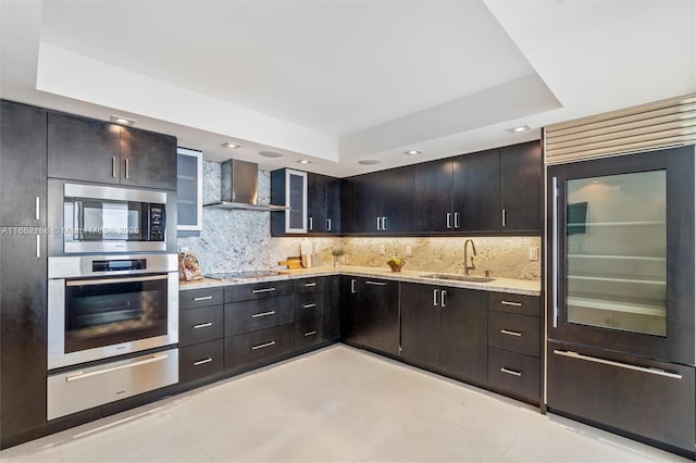 kitchen featuring sink, built in appliances, a raised ceiling, wall chimney range hood, and backsplash