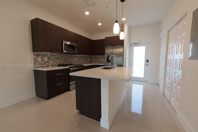 kitchen featuring appliances with stainless steel finishes, decorative light fixtures, decorative backsplash, dark brown cabinetry, and a center island with sink