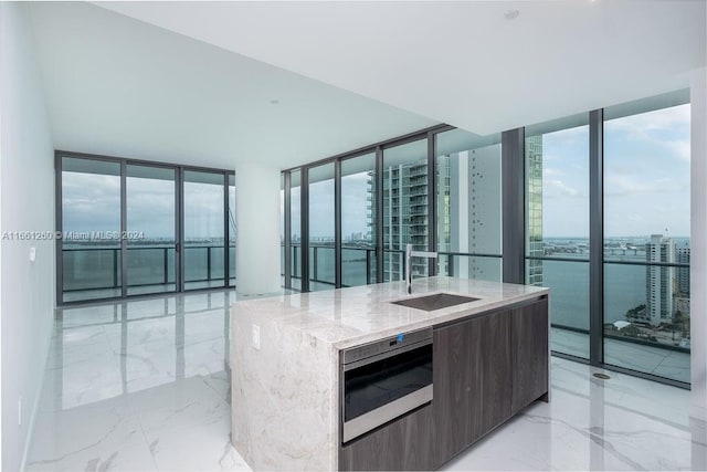 kitchen with a wall of windows, a kitchen island with sink, light stone countertops, and a wealth of natural light