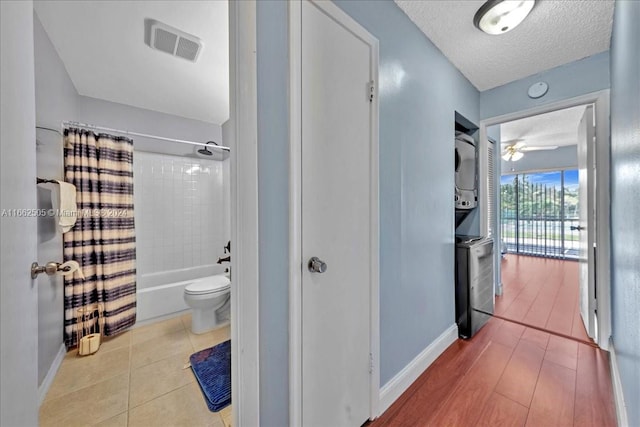 bathroom with stacked washer / drying machine, toilet, shower / bath combination with curtain, hardwood / wood-style floors, and ceiling fan