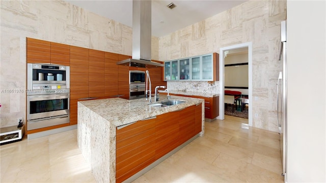 kitchen with tile walls, island range hood, light stone countertops, a kitchen island with sink, and sink