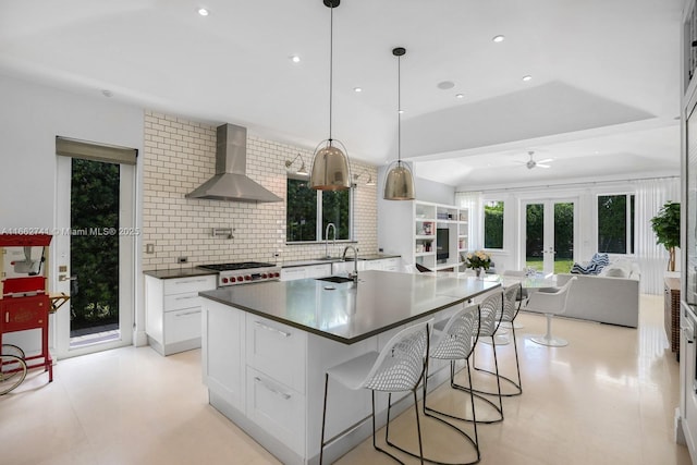 kitchen with dark countertops, backsplash, open floor plan, a kitchen island with sink, and wall chimney range hood