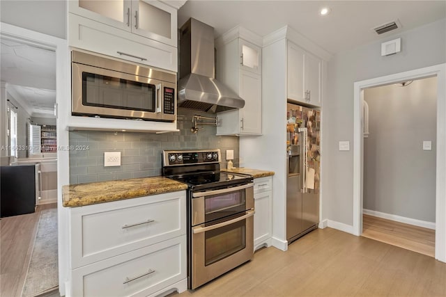 kitchen featuring wall chimney range hood, white cabinetry, decorative backsplash, appliances with stainless steel finishes, and light wood-style floors