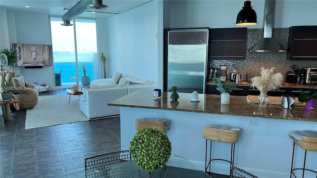 kitchen with stainless steel built in fridge, hanging light fixtures, backsplash, ventilation hood, and dark stone countertops