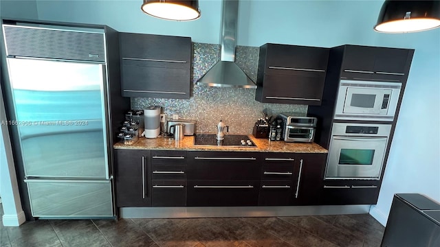 kitchen featuring dark tile patterned flooring, built in appliances, wall chimney exhaust hood, backsplash, and light stone countertops