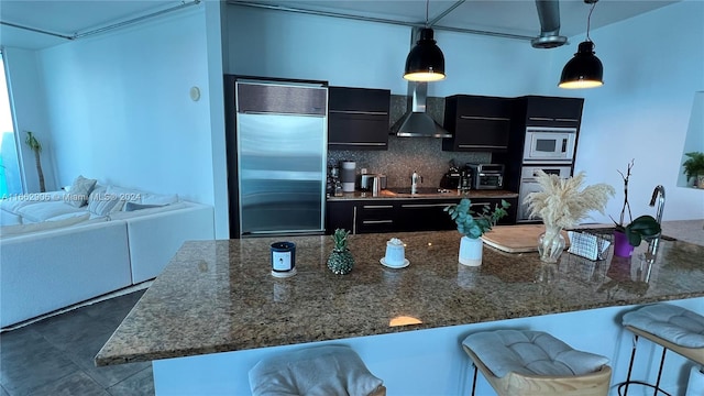 kitchen with hanging light fixtures, a breakfast bar area, built in appliances, and wall chimney range hood