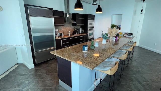 kitchen with a kitchen bar, wall chimney exhaust hood, a center island with sink, appliances with stainless steel finishes, and decorative light fixtures