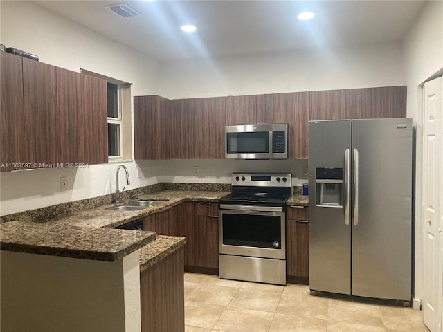 kitchen featuring kitchen peninsula, appliances with stainless steel finishes, dark stone counters, dark brown cabinets, and sink