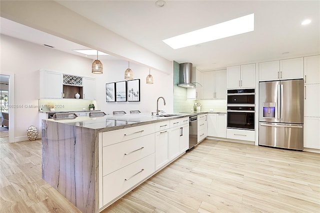 kitchen featuring a skylight, stainless steel appliances, wall chimney range hood, kitchen peninsula, and decorative light fixtures